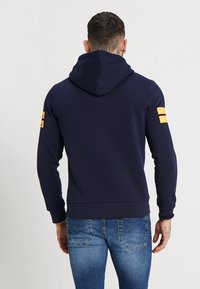 Sudadera navy de tejido suave con una gran capucha, que presenta rayas amarillas en la manga izquierda y un diseño ajustado en la cintura y los puños.