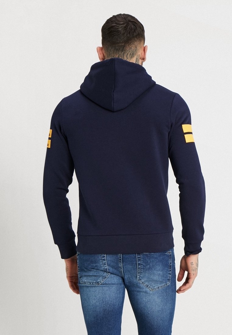 Sudadera navy de tejido suave con una gran capucha, que presenta rayas amarillas en la manga izquierda y un diseño ajustado en la cintura y los puños.