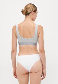 Woman shown from behind wearing a light gray sports bra and white underwear, with hair styled in a braided bun against a plain background.