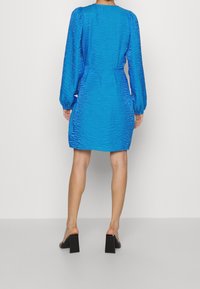 Mujer vistiendo un vestido mini de manga larga texturizado en un brillante color azul, con mangas fruncidas y zapatos de tacón bloque negro, vista desde la parte posterior.