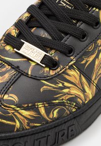 Svart sneaker med ett blommönster i gult och orange, med en texturerad läderöverdel, svarta snören och ett guldemblem med texten "VERSACE JEANS COUTURE."