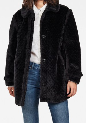 Femme portant un manteau noir à texture pelucheuse par-dessus une chemise blanche boutonnée et un jean bleu, debout devant un fond blanc.