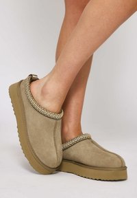 Zoccoli beige slip-on realizzati in morbido suede, con un bordo decorativo intrecciato e suola in gomma caratterizzata da un motivo a zig-zag.