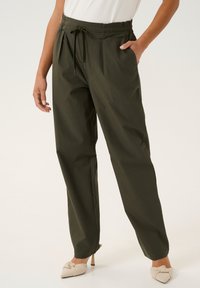 Pantaloni verde oliva, leggeri, con vita elastica e cordoncino. Presentano tasche laterali e un design a gamba larga. Abbinati a mule beige annodati.