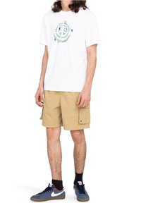 T-shirt blanc avec un motif graphique multicolore sur le devant, associé à un short cargo beige avec poches latérales, porté avec des baskets bleues.