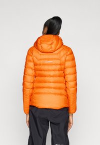 Oranžs pūkains jaku ar pārbaudītu tekstūru un kapuci. Iezīmē redzamās šuves un "Mammut" logo augšējā aizmugurē.