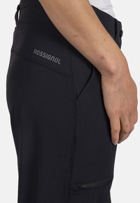 Czarne spodnie outdoorowe o gładkiej fakturze, z kieszenią zapinaną na zamek oraz logo na pasie, zaprojektowane z myślą o elastyczności i komforcie.