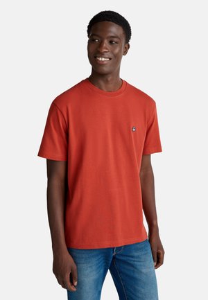 Red cotton t-shirt with a round neckline and short sleeves; features a small blue logo on the left chest; worn with blue jeans.