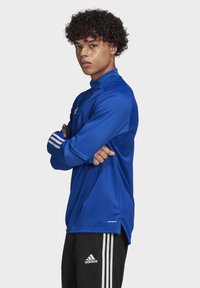 Blå atletisk langærmet top med høj hals, tekstureret stof og hvide tre-stribe accenter påærmerne; parret med sorte bukser med striber.