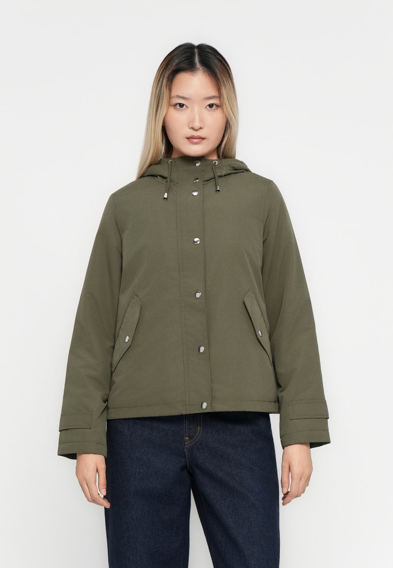Femme de face portant une veste à capuche olive boutonnée et un jean bleu foncé sur fond blanc.