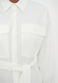 Chemise blanche à manches longues avec col pointu, fermeture boutonnée devant, deux poches poitrine boutonnées et ceinture nouée à la taille.
