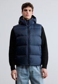 Colete acolchoado azul-marinho com capuz, fecho de correr e secções acolchoadas. Vestido sobre um suéter preto de malha, combinado com jeans azul-claro.