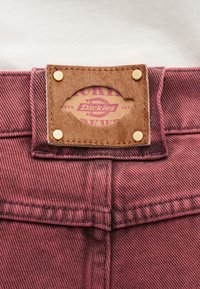 Καφέ δερμάτινο λογότυπο Dickies με μεταλλικά πριτσίνια στη μέση κόκκινου τζιν υφάσματος.