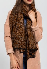 Femme portant une écharpe épaisse marron et noire à texture, par-dessus un manteau rose clair aux manches retroussées, tenant l'écharpe près de son cou.