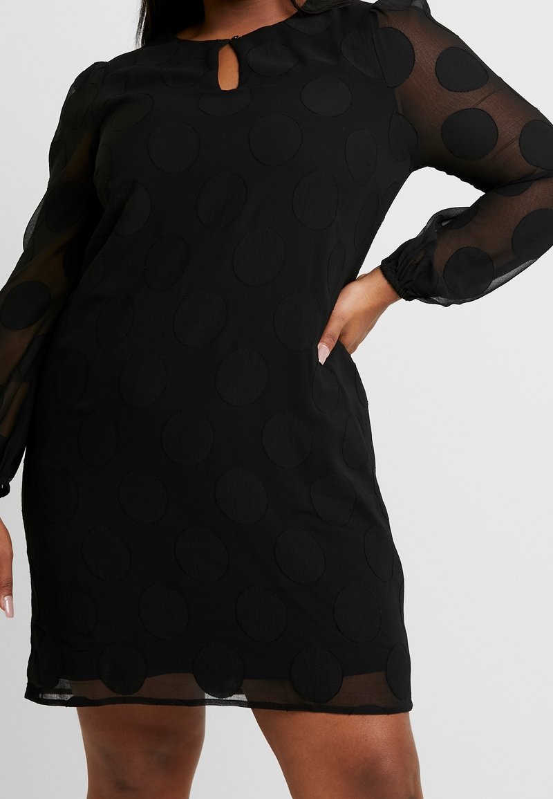 Robe noire avec de grands pois texturés, manches longues transparentes et un petit col en forme de goutte portée par une personne avec la main sur la hanche.