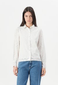 White long-sleeve shirt with a button front, featuring eyelet embroidery and a slightly structured design. Paired with high-waisted blue jeans.