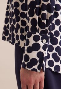 Camicia a maniche lunghe in tessuto bianco con grandi pois blu navy, con dettaglio a bottone sul polsino e leggero plissettato vicino alla cucitura.