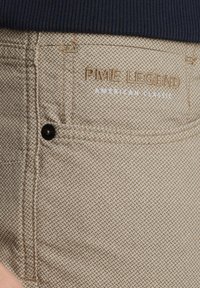 Pantalons texturés beiges avec un motif en losange, dotés d'un bouton noir et d'un logo brodé portant l'inscription "PME LEGEND AMERICAN CLASSIC."