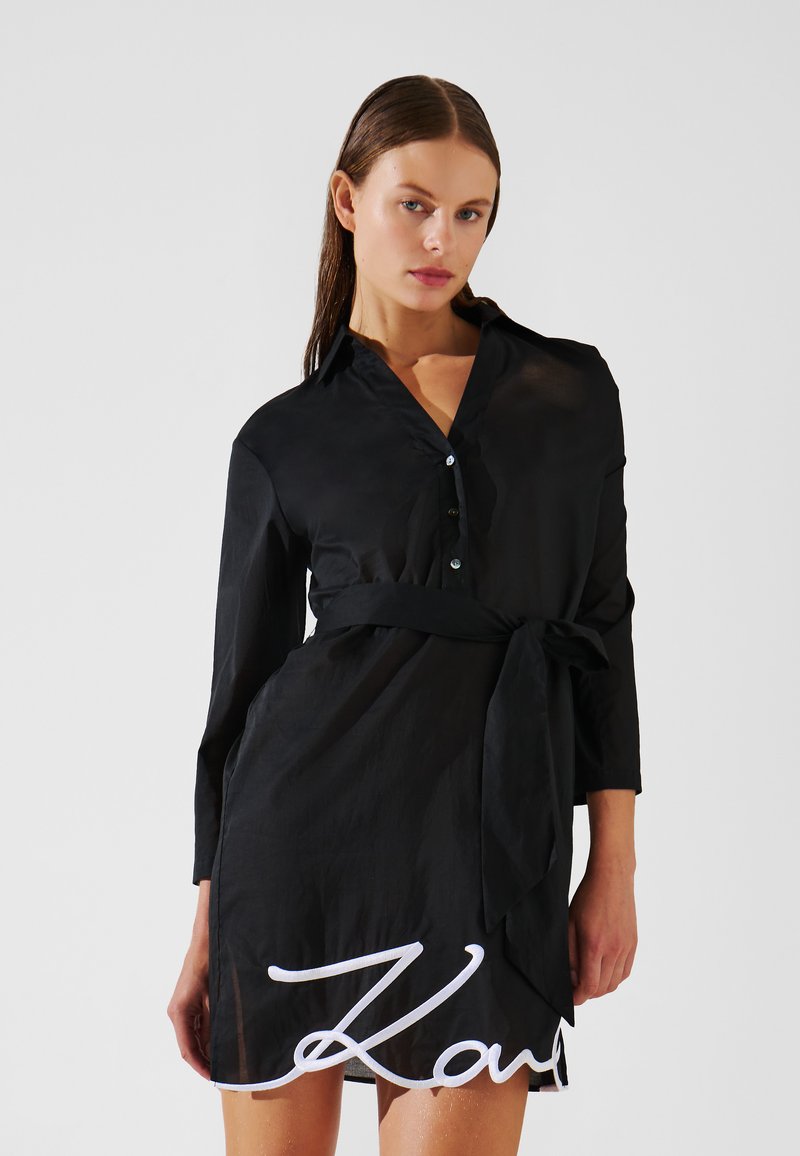 Vestido negro de camisa hecho de una tela ligera, que presenta un collar, mangas largas, cintura ajustada y un detalle de nombre bordado en blanco en el dobladillo.
