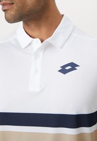 Hvid polo skjorte lavet af tekstureret stof, med et marineblåt logo og vandrette striber i marineblå og beige hen over forsiden.
