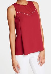 Top senza maniche rosso con scollatura trasparente, caratterizzato da dettagli con borchie argentate lungo un design a zig-zag e una vestibilità rilassata.