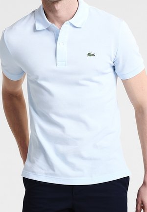 Homme portant un polo bleu clair à manches courtes avec un petit logo crocodile vert sur la poitrine gauche.