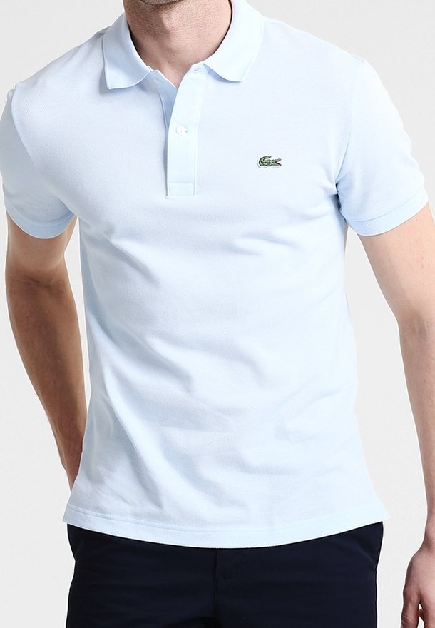 Homme portant un polo bleu clair à manches courtes avec un petit logo crocodile vert sur la poitrine gauche.
