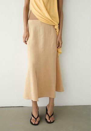 Kobieta w beżowej spódnicy midi z bocznym rozcięciem, żółtej bluzce związanej w pasie i czarnych sandałach na obcasie, stojąca przy białej ścianie.