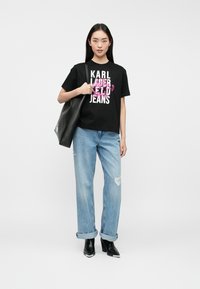 T-shirt de algodão preta com texto gráfico em branco e rosa, combinada com jeans de ganga azul claro desgastados e botins pretos de salto.