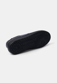 Black sneaker with a rubber sole featuring a herringbone pattern and circular accent details. Smooth upper with stitched seams.