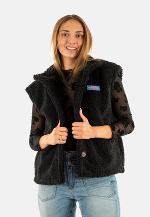 Gilet in pelliccia sintetica nera con cappuccio, con chiusura frontale a pulsante e un patch colorato del marchio. Indossato su una blusa nera trasparente con stampa floreale.
