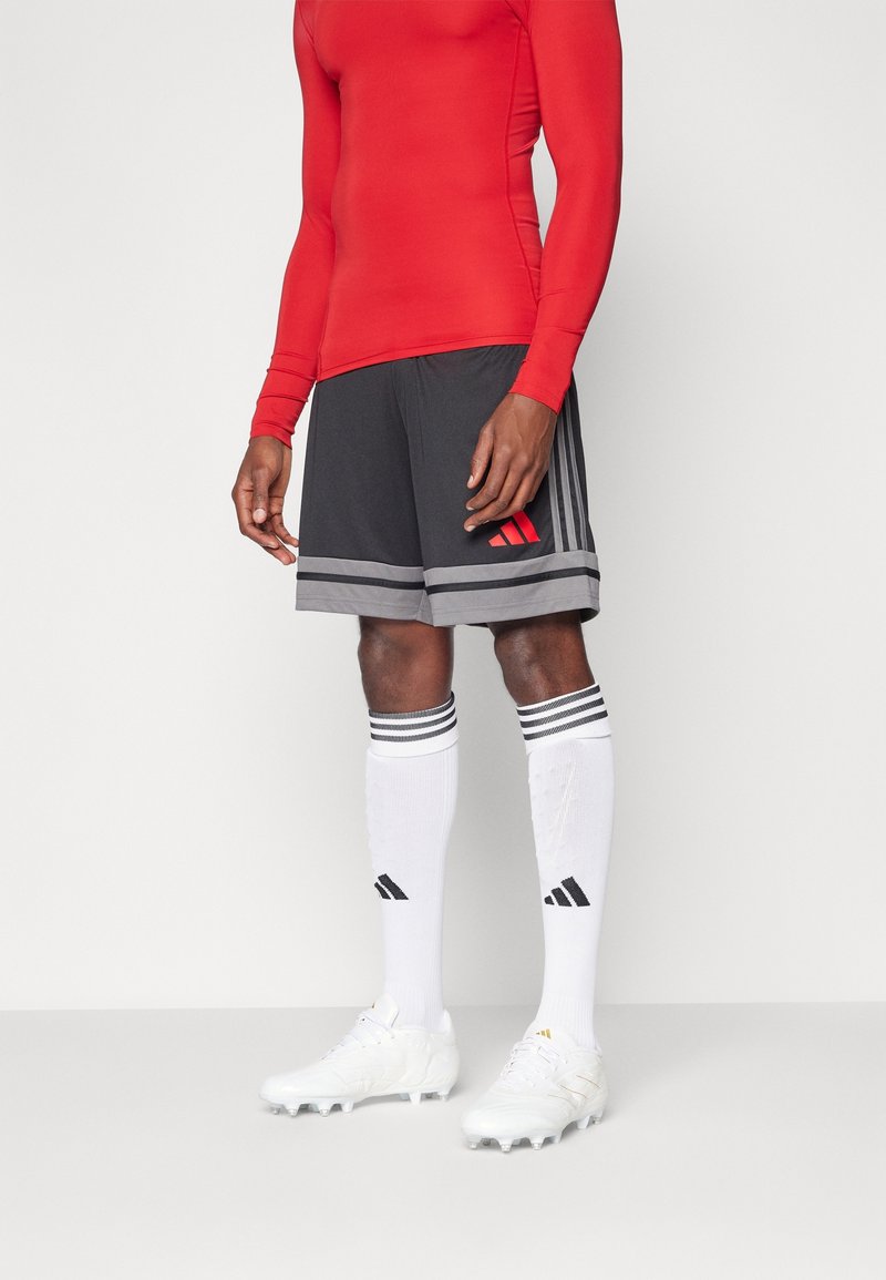 Personne portant un t-shirt rouge à manches longues, un short de sport noir et gris, des chaussettes blanches hautes avec des rayures noires et des crampons blancs, se tenant sur un fond uni.