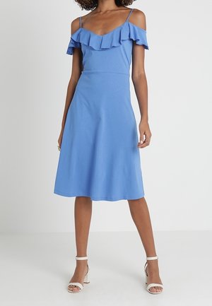 Niebieska sukienka bez ramiączek z falbaną przy dekolcie i rozkloszowanym fasonie, wykonana z lekkiego materiału. W zestawieniu z białymi sandałami na obcasie.