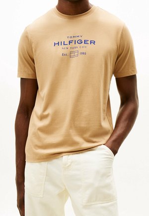Mężczyzna w beżowej koszulce Tommy Hilfiger z niebieskim logo i jasnych spodniach, stoi z jedną ręką w kieszeni.