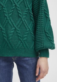 Pull en maille vert avec un motif en torsade et des manches texturées, doté d'un ourlet et de poignets côtelés. Associé à un jean en denim bleu.