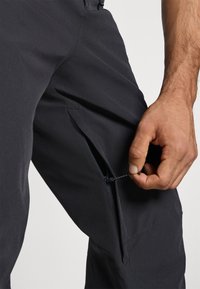 Črne hlače iz lahkotnega materiala z zadrgo na ventilaciji; roka prilagaja vrvico na pasu.