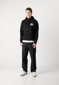 Sudadera negra con capucha, bolsillo frontal y diseño gráfico. Combinada con jeans de mezclilla oscura y zapatillas blancas. Textura suave en general.