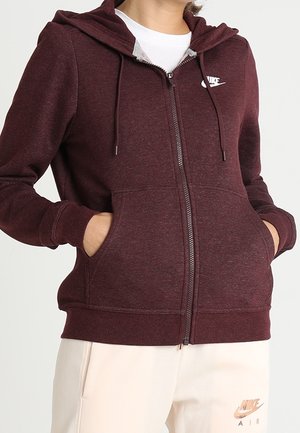 Person wearing a maroon zip-up Nike hoodie with hands in front pockets, paired with light-colored Nike Air pants.