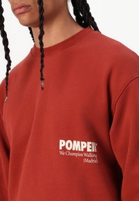 Rød hettegenser laget av bomull, med ribbestrikket halslinning. Forsiden har hvit tekst: "POMPEII We Champion Walking (Madrid)."