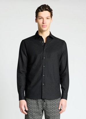 Junger Mann mit kurzen lockigen Haaren, der ein schwarzes Hemd mit Knöpfen und schwarz-weiß gemusterte Hose trägt, steht vor einem einfarbigen hellen Hintergrund.