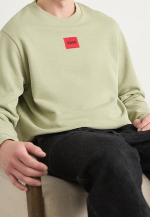 Person trägt einen hellgrünen Pullover mit einem roten "HUGO"-Aufnäher auf der Brust und sitzt mit den Händen auf den Beinen.