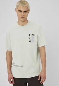 T-shirt en coton vert clair avec des manches courtes, présentant un design graphique noir et lavande avec l'inscription "Piña Colada" et des accents de texte.