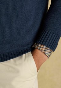 Mees, kellel on seljas tumesinine kampsun triibulise särgi üle, käsi kreemivärvises pükste taskus neutraalse tausta ees.