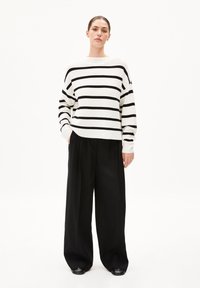 Gestreifter Pullover in Weiß und Schwarz, kastiger Schnitt mit weiten Ärmeln. Kombiniert mit lockeren schwarzen Hosen und schwarzen Schuhen. Minimalistischer Stil.