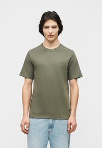 Νεαρός άνδρας που στέκεται με το πρόσωπο προς τα εμπρός, φορώντας κοντομάνικο χακί πράσινο μπλουζάκι και ανοιχτόχρωμο τζιν, μπροστά σε απλό φόντο.