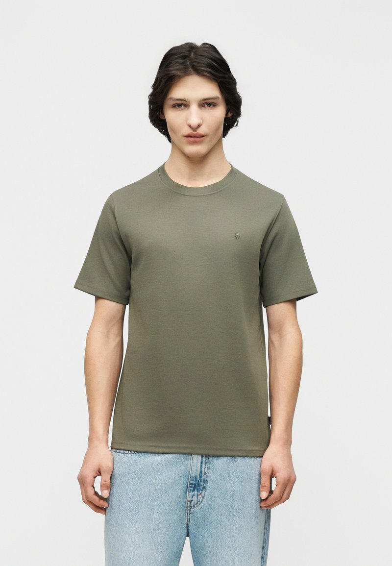 Νεαρός άνδρας που στέκεται με το πρόσωπο προς τα εμπρός, φορώντας κοντομάνικο χακί πράσινο μπλουζάκι και ανοιχτόχρωμο τζιν, μπροστά σε απλό φόντο.