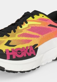 Żółty i różowy but do biegania z czarnymi sznurowadłami i podeszwą, biała midsole i dużym różowym logo HOKA po boku.