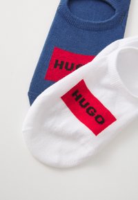 Két pár bokazokni: az egyik sötétkék, a másik fehér. Mindkettőn piros címke található, rajta fekete "HUGO" felirattal. Pamut keverék anyag, könnyű kialakítás.