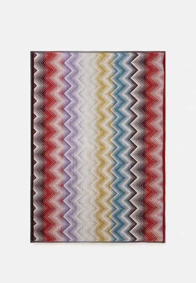 Tapis rectangulaire avec des rayures en zigzag horizontales rouge, violet, blanc, jaune, bleu et noir sur fond blanc.