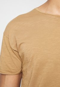 Tričko s krátkým rukávem a kulatým výstřihem v světle hnědé barvě, vyrobené z měkké, texturované látky s mírně volnějším střihem a jemným vertikálním vzorem.